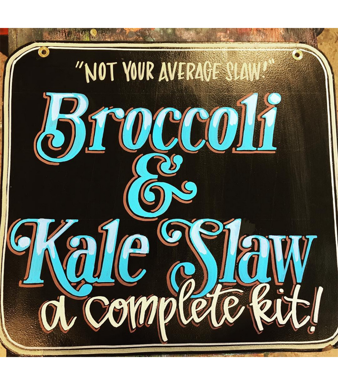 A hand-painted sign with vibrant, ornate lettering that reads 'Broccoli & Kale Slaw' in large blue and white text with red shadows. Above the main text, the phrase 'Not Your Average Slaw' is written in smaller white letters. Below, in cursive script, it says 'a complete kit!' The sign has a decorative border and is set against a black background, giving it a classic yet eye-catching appearance, perfect for a market or restaurant