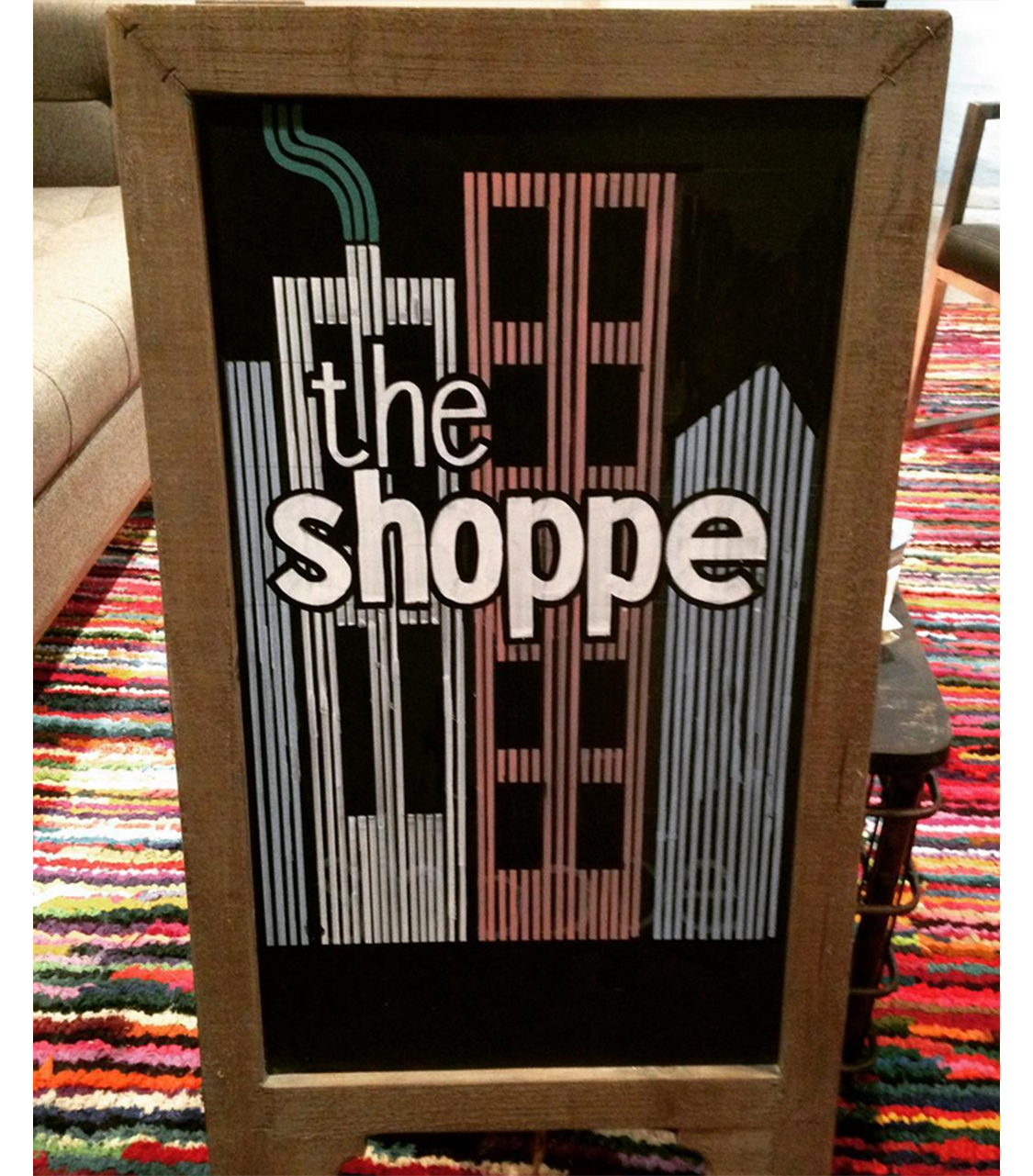 A wooden-framed signboard displaying a hand-painted design with the words 'The Shoppe' in bold, white letters. The background features a stylized illustration of tall buildings in muted shades of blue and pink, with a green smoke stack on the left. The sign has a vintage, urban feel, blending modern typography with a retro architectural motif, placed on a colorful, striped rug.